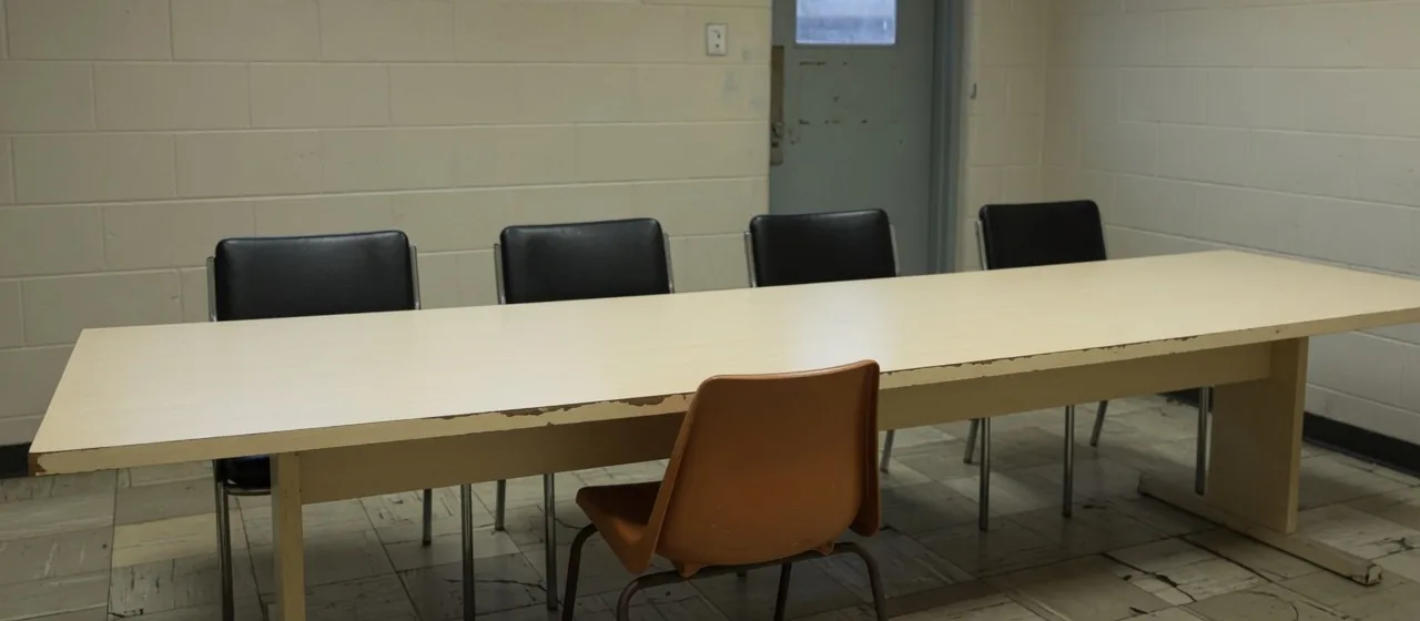 An empty institutional hearing room: a long table with four vacant chairs behind it and one lone chair pulled up alone on the near side