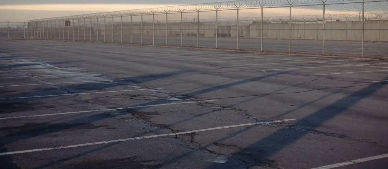 An empty prison parking lot at dawn, cracked asphalt running to a razor-wire fence with low buildings beyond it
